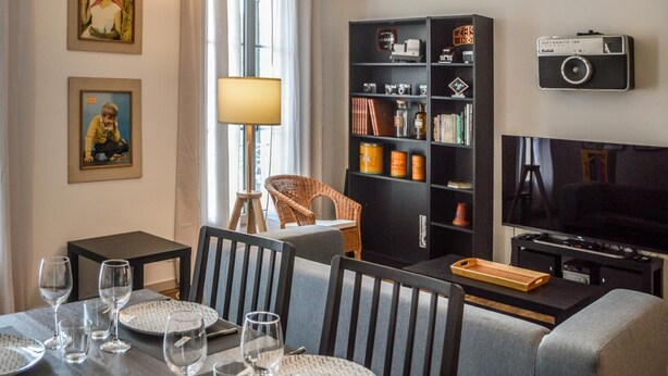 A dining area with a table set for four, a television, and a bookshelf with various items.