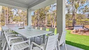 A covered outdoor dining area with a long white table and matching chairs, surrounded by a natural landscape.