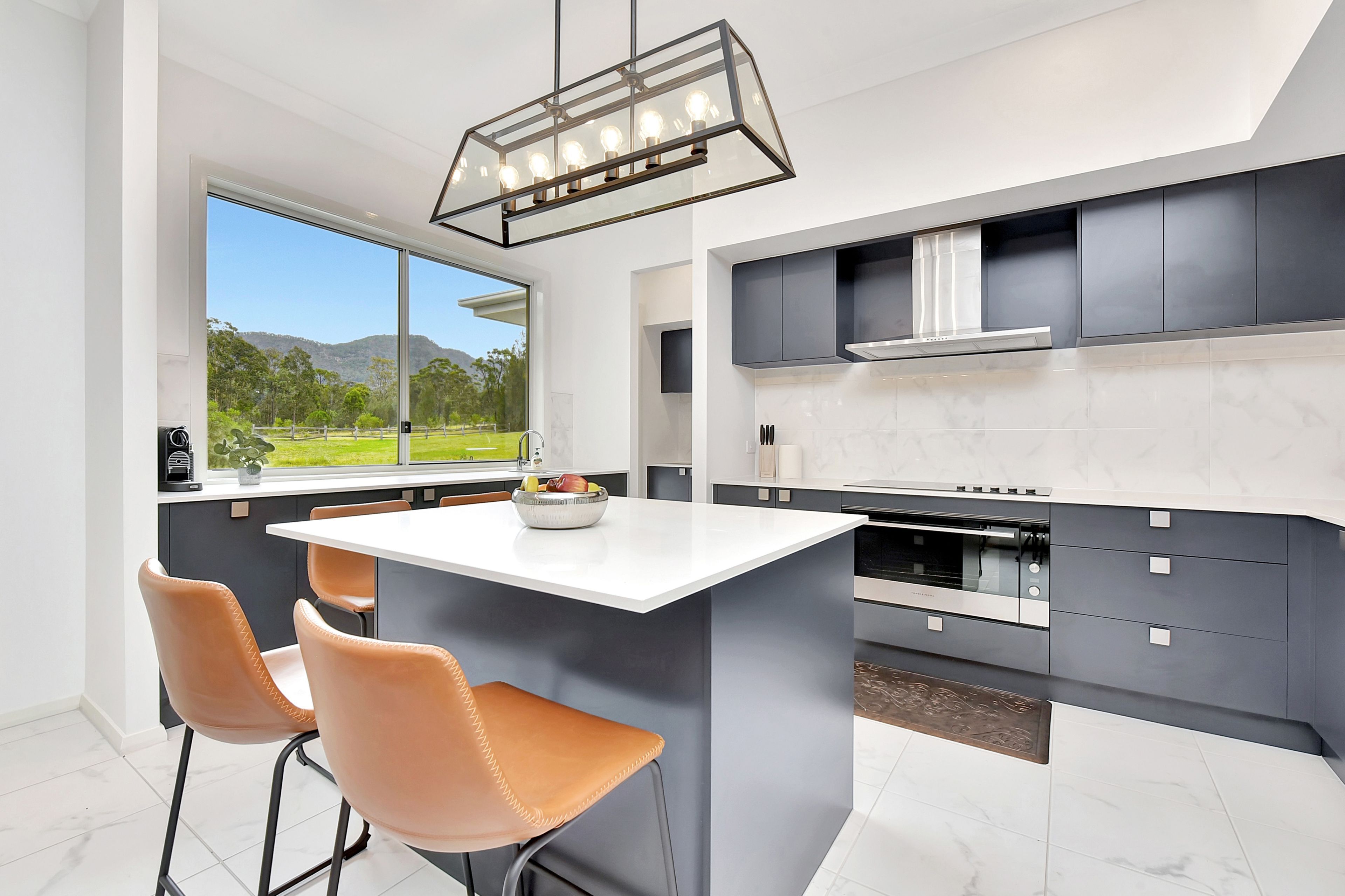 A modern kitchen with a central island, two chairs, a window with a view of greenery, and a pendant light fixture.