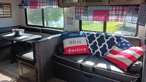Interior d'una caravana acollidora amb zona de menjador, espai d'estar amb coixins de temàtica americana i finestra amb vista a vegetació.
