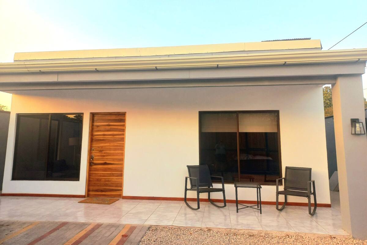 A modern building with a flat roof, large windows, and a door with a wooden panel. There are two chairs outside.