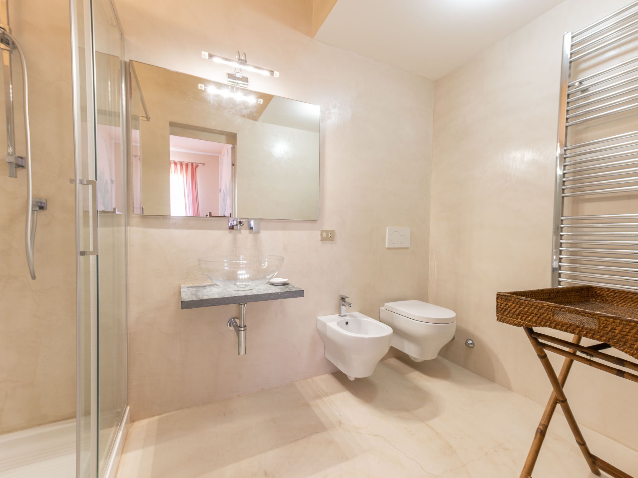 Ein modernes Badezimmer mit einer Duschkabine aus Glas, einem Waschbecken aus Glas, einem Bidet und einem Korbwägelchen auf einem Hocker.