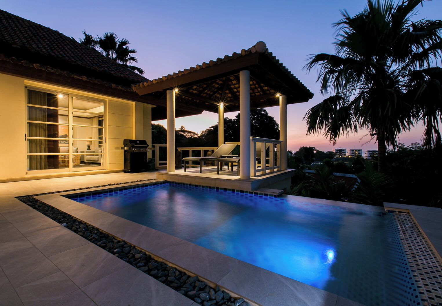 A swimming pool with a pergola and outdoor seating, surrounded by a house with large windows and a grill.