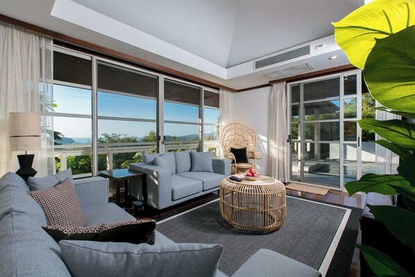 A modern living room with a grey sofa, a wicker coffee table, and a view of a pool and greenery.