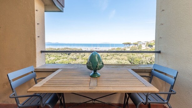A wooden table with two chairs and a green vase on a balcony overlooking a coastal area with a view of the sea and distant buildings. - I Boboli by Interhome (Punta Ala)
