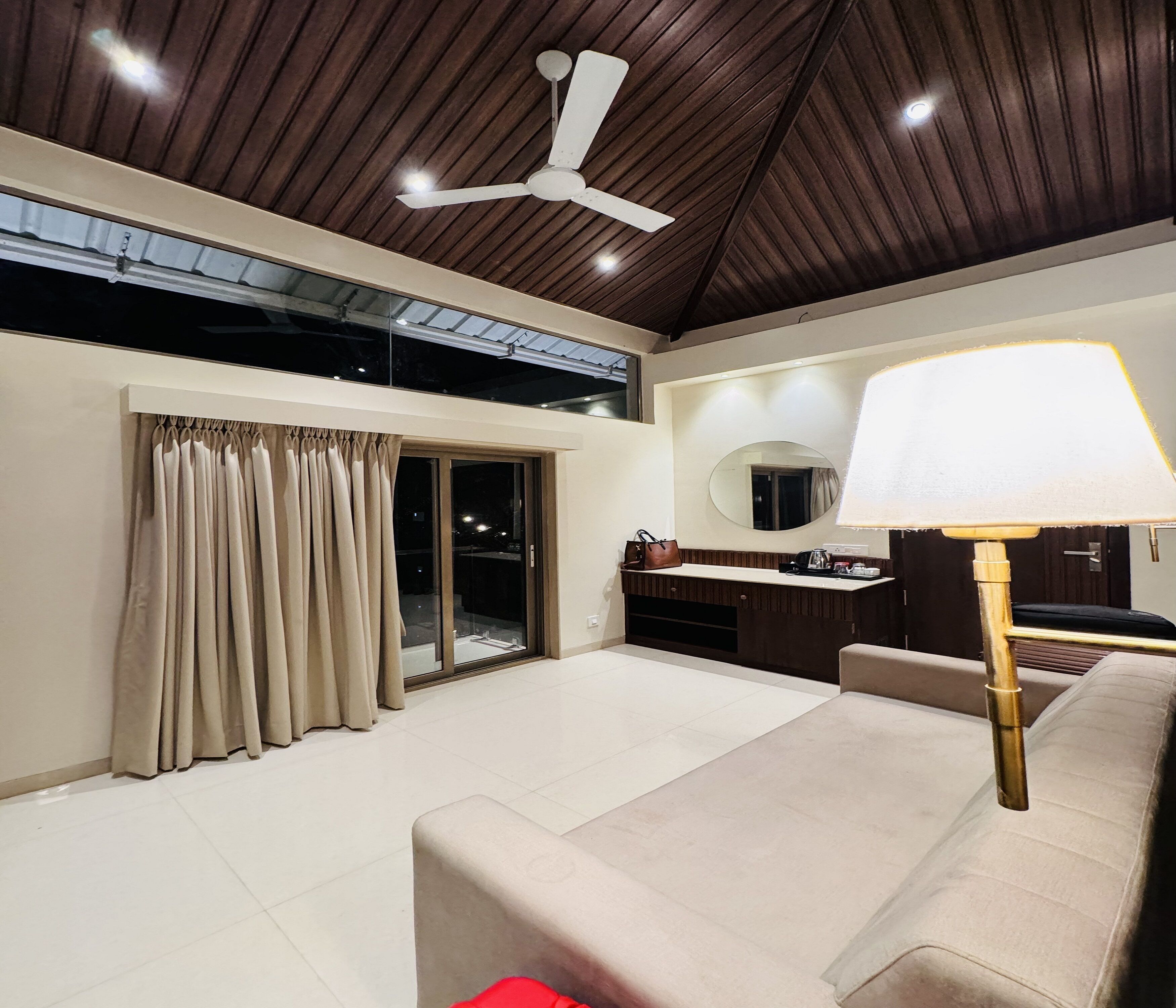 A modern hotel room with a ceiling fan, a sofa, a lamp, a sliding glass door, and a small kitchenette.