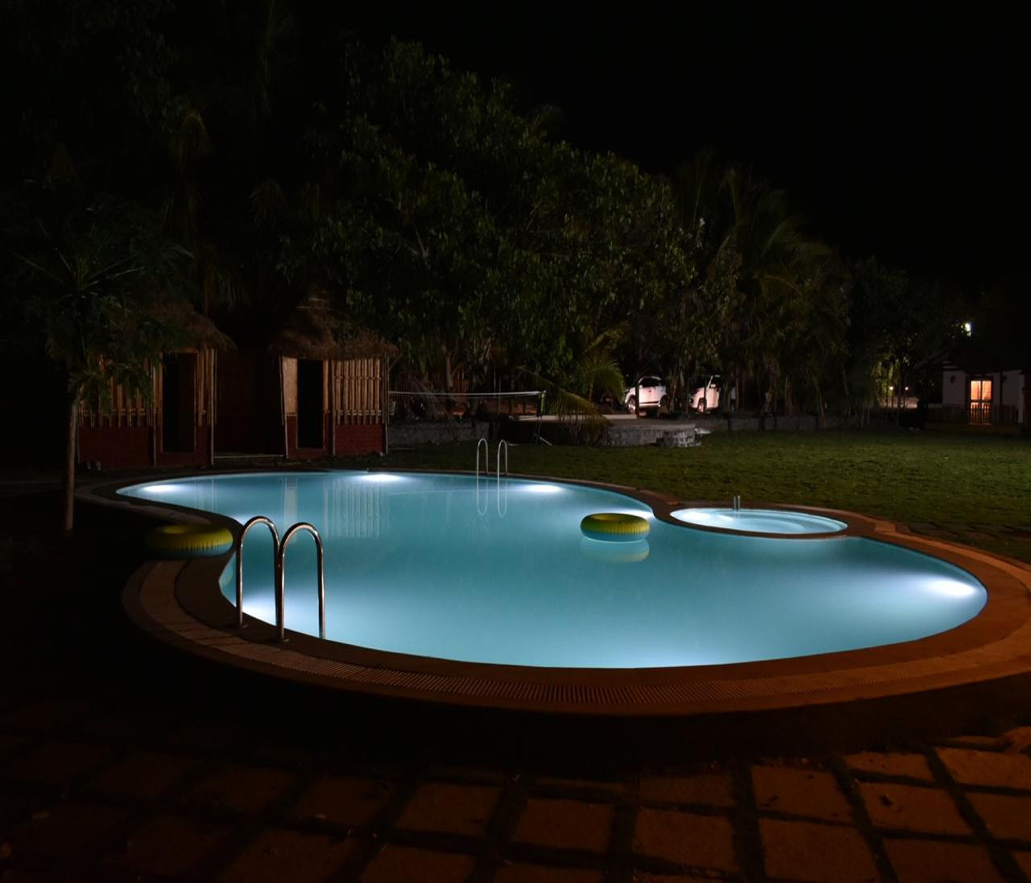 Una piscina nocturna con un muelle de madera y una estructura de techo de paja al fondo.