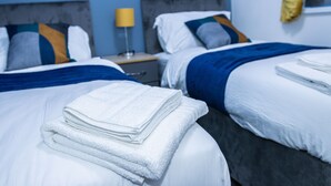 A hotel room with two beds, each with white bedding and blue accents, a nightstand with a lamp, and a window with yellow curtains. - Impeccable 4-Bed House in Telford, West Midlands (Telford)
