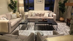 Un salon avec un ensemble de canapés de couleur beige, une table basse et un tapis. Il y a deux fenêtres avec des rideaux, et une plante est visible sur le côté droit.