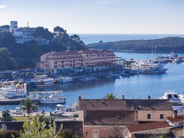 Un port marítim costaner amb nombroses embarcacions amarrades, un gran edifici d'hotel i vista al mar i a les colines al fons.