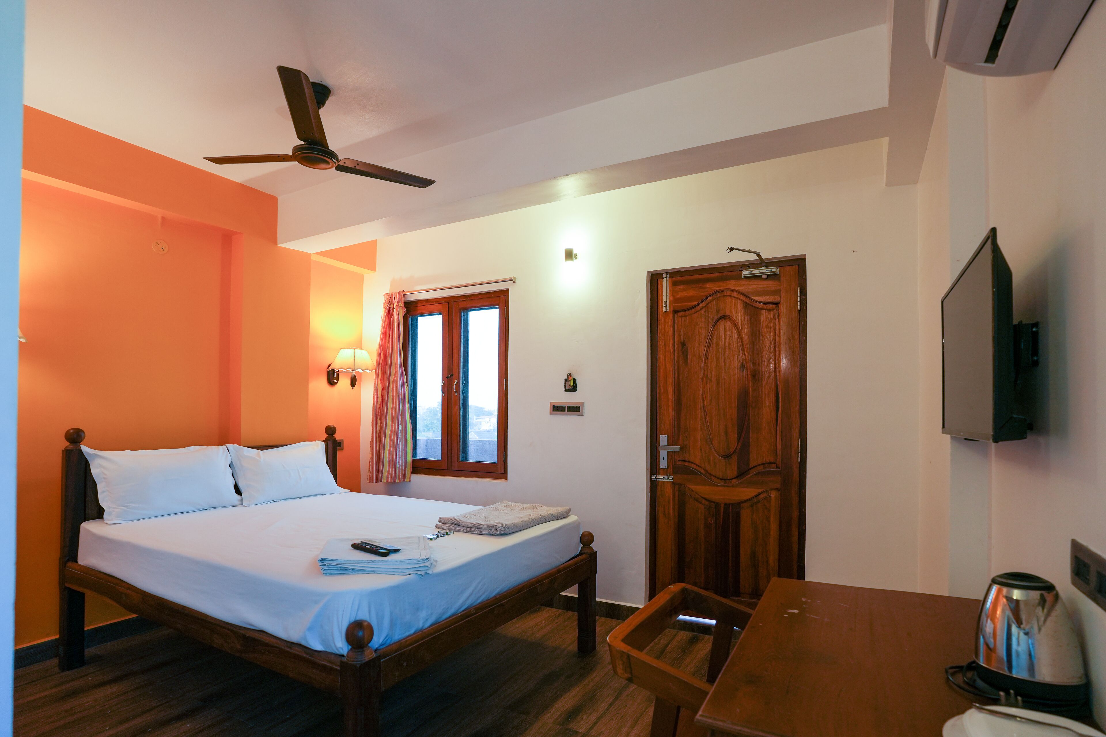 A bedroom with a wooden bed, a ceiling fan, a desk with a kettle, and a television on the wall.