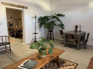 A living room with a wooden table, a potted plant, a bookshelf, and a dining area in the background. - Adobe Shotgun House (Tucson)