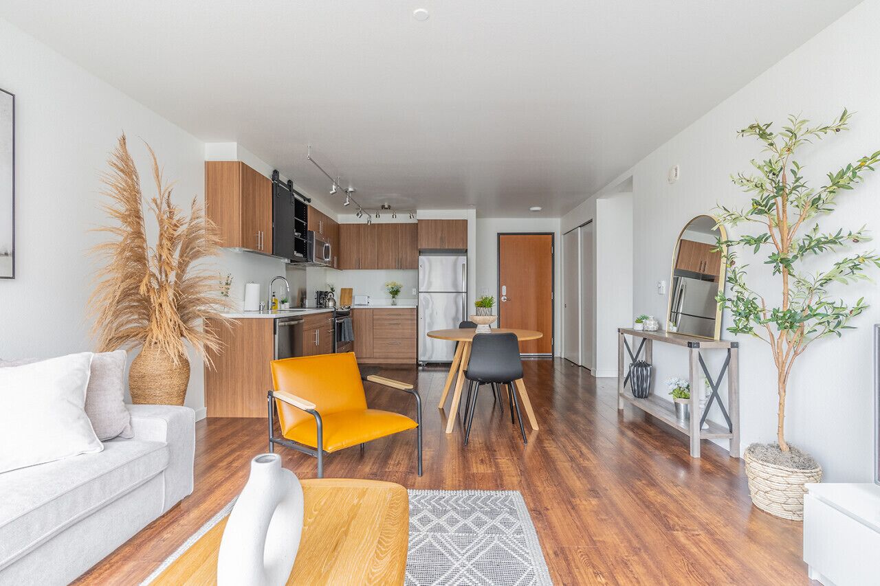 Una sala de estar moderna con un sofá, una mesa de comedor con sillas, una zona de cocina con armarios y electrodomésticos, y una planta en maceta.