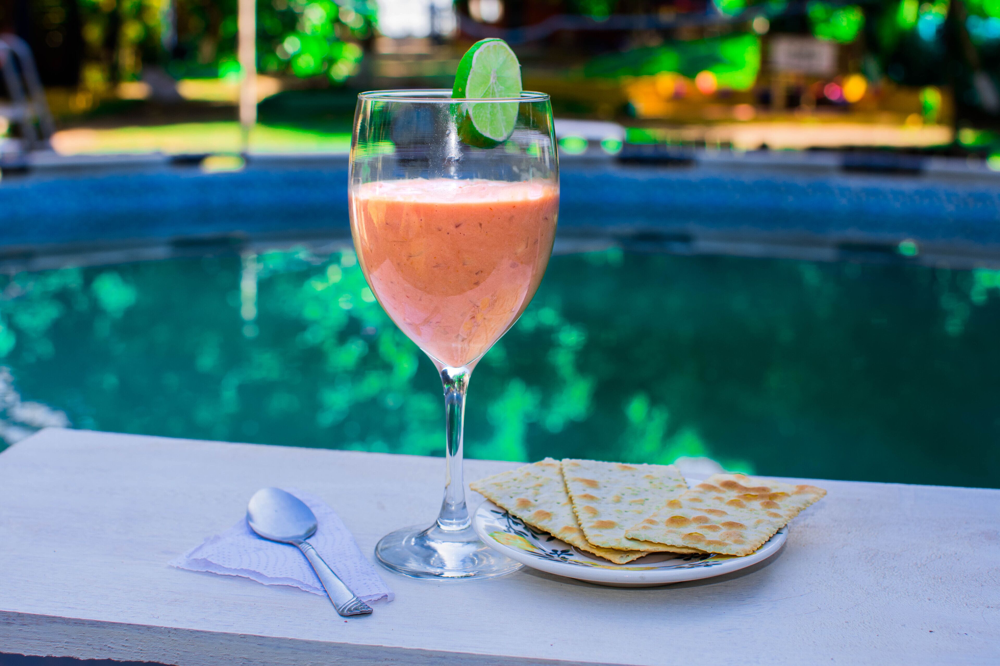 Un vaso de bebida rosada con una rodaja de lima, una cuchara y un plato de pan plano junto a una mesa al borde de la piscina.