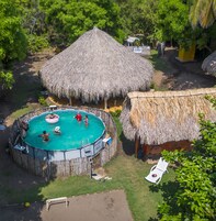 Una piscina circular con techo de paja, rodeada por una valla de madera, y una pequeña cabaña con techo similar cerca.