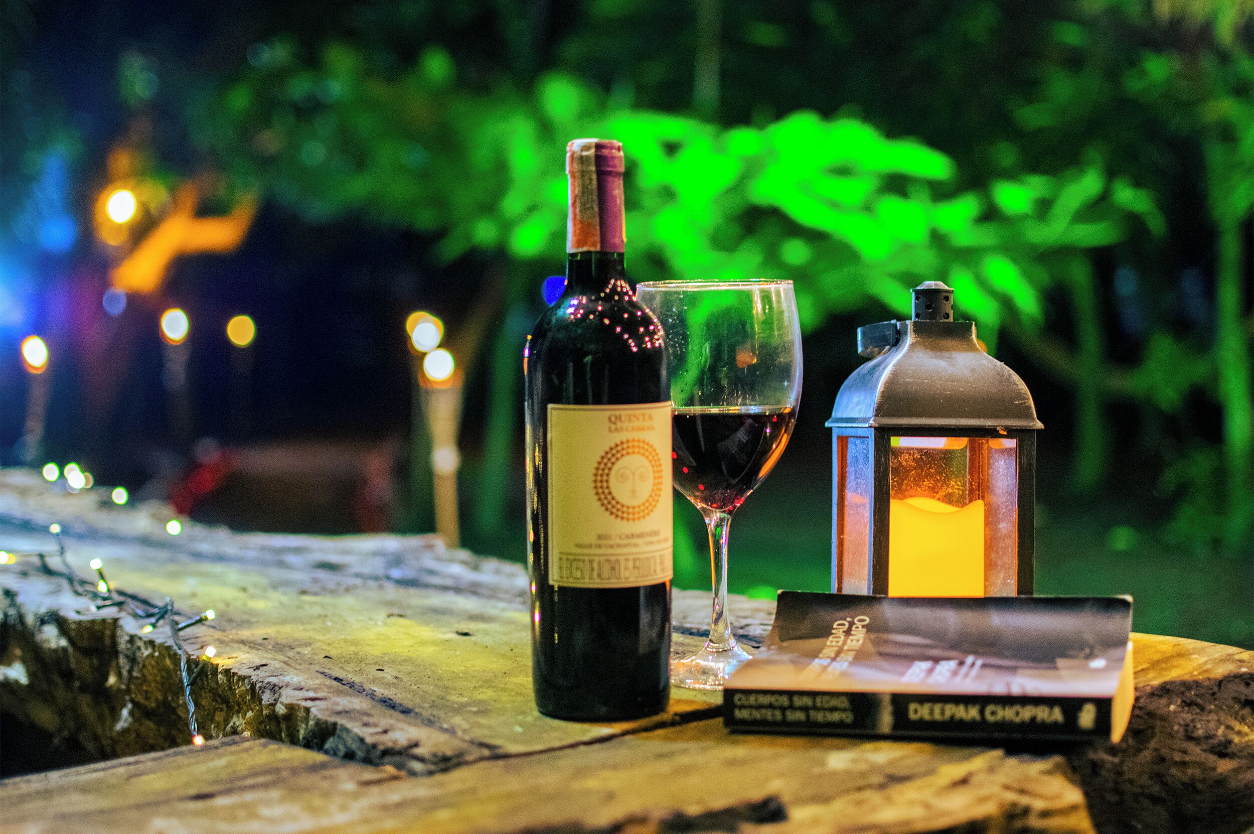 Una botella de vino, una copa de vino y un libro sobre una superficie de madera con un farol al fondo.