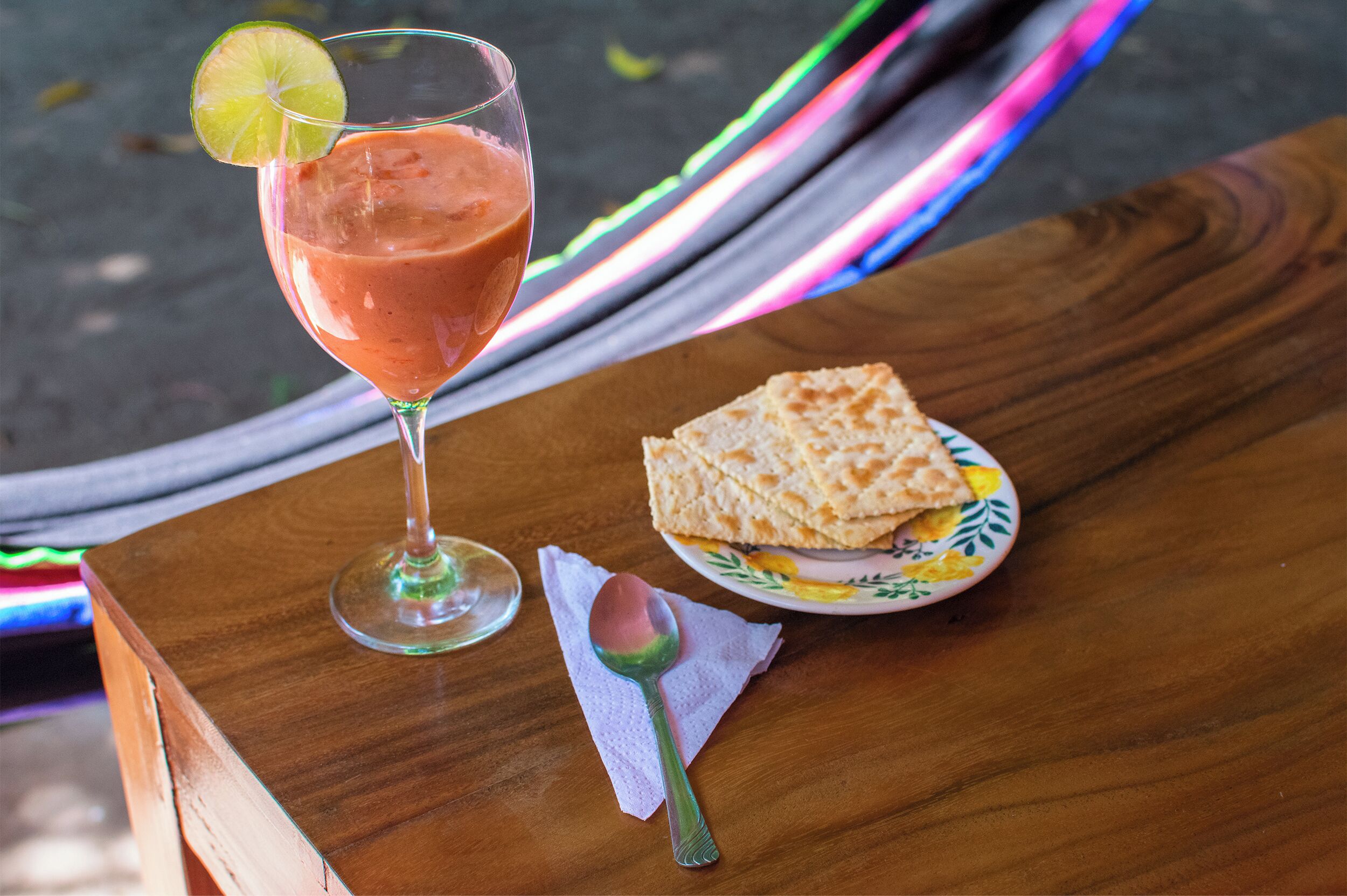 A glass of pink drink with a lime slice on a wooden table, accompanied by a plate of flatbread and a spoon.