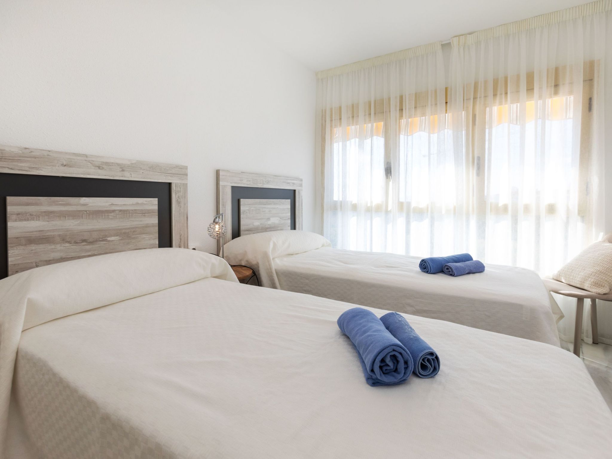 Una camera d'albergo con due letti, un comodino, una lampada e una finestra con tende leggere.