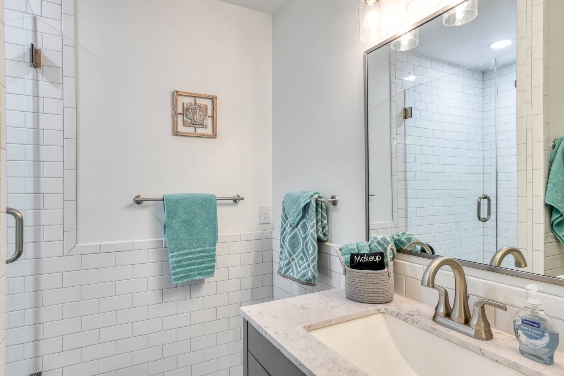 Un baño con azulejos blancos tipo metro, cerramiento de ducha de vidrio, lavamanos con encimera de mármol, espejo y toallero con varias toallas.
