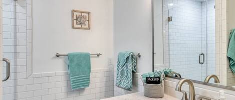Un baño con azulejos blancos tipo metro, cerramiento de ducha de vidrio, lavamanos con encimera de mármol, espejo y toallero con varias toallas.