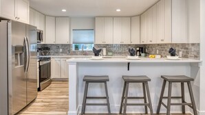 A modern kitchen with white cabinets, a stainless steel refrigerator, a large island with stools, and a window with blinds. - Maritime Place Unit A (St. Augustine)