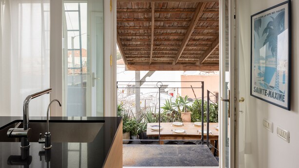 Ein modernes Badezimmer mit schwarzem Waschtisch und einer Duschkabine. Eine Holzbalkendecke und eine Tür zu einem Balkon mit Pflanzen und einem Tisch sind ebenfalls vorhanden.