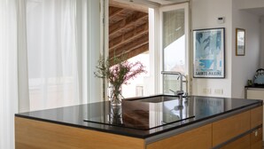 Eine Küche mit schwarzem Arbeitsplatz, Holzschränken, einer Edelstahl-Spüle und einem Fenster, durch dessen offene Tür eine Holzbalkendecke sichtbar ist.