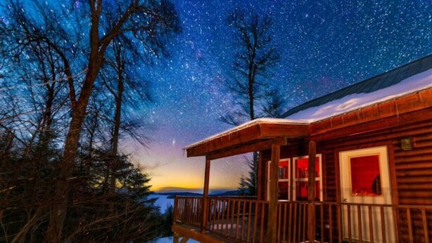 Una cabaña de madera con balcón que da a un paisaje nevado y un cielo nocturno estrellado.