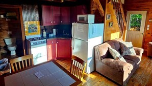 Interior acogedor de una cabaña, con una zona de cocina que incluye estufa, microondas y refrigerador. Hay una mesa de comedor con sillas y un sofá con cojines. Una escalera de madera conduce a un nivel superior.