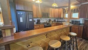 A kitchen with wooden cabinets, a stainless steel refrigerator, a stove, and a bar area with stools. - Woods All Around-Direct ATV*Hot Tub*Beach Access*A/C*Pool Table*Washer/Dryer (Beaver Cove)