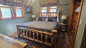 A rustic bedroom with a wooden bed frame, two bedside tables with lamps, a wooden desk, and a window with curtains. - Woods All Around-Direct ATV*Hot Tub*Beach Access*A/C*Pool Table*Washer/Dryer (Beaver Cove)