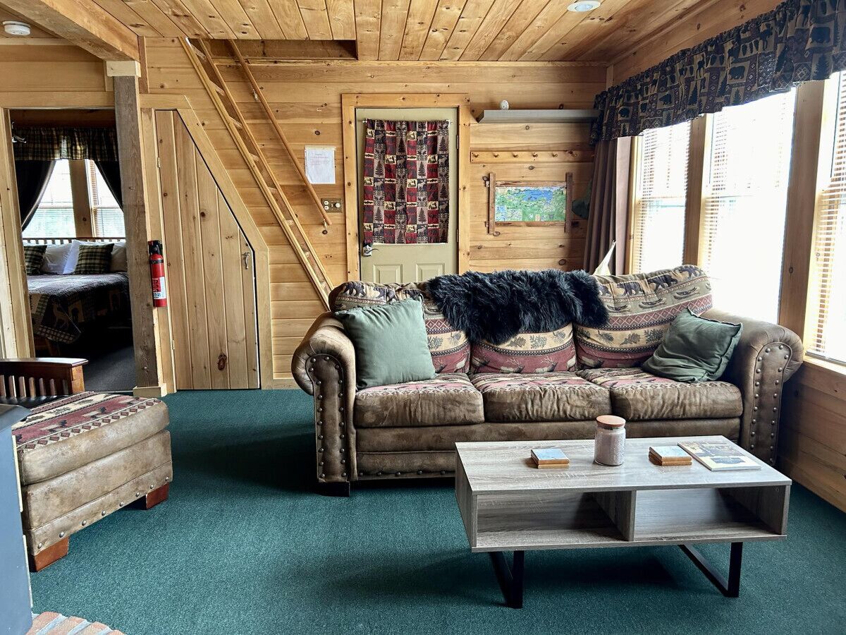 A cozy cabin interior with a wooden staircase, a patterned couch, a small table with books and candles, and a rustic wooden floor.