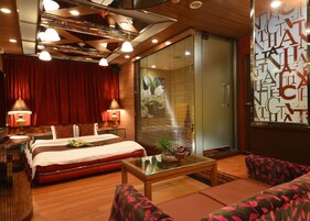 Una habitación de hotel con cama, dos lámparas de noche, una mesita de vidrio, un sofá y una puerta corrediza de cristal con persianas.