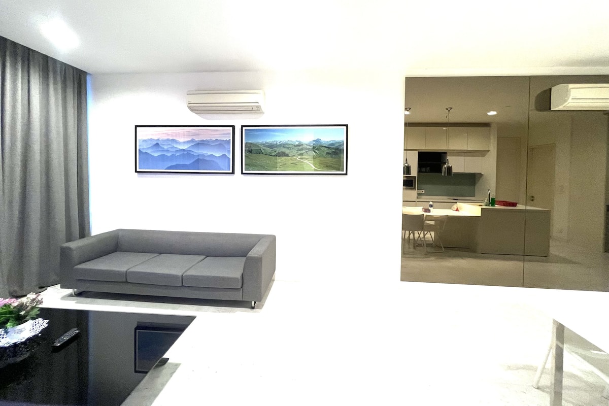 A modern living room with a grey sofa, a flat-screen TV, and a coffee table. There are two framed landscape paintings on the wall.