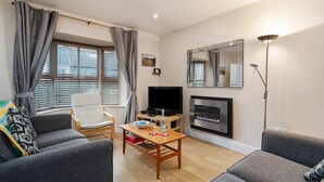 A living room with a sofa, armchair, TV, fireplace, and a table with a bowl and wine glasses. - Park Place Court 2 - Close to Beach (Tenby)