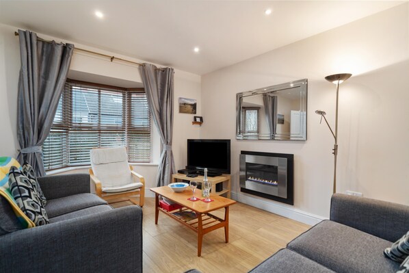 A living room with a sofa, armchair, TV, fireplace, and a table with a bowl and wine glasses. - Park Place Court 2 - Close to Beach (Tenby)
