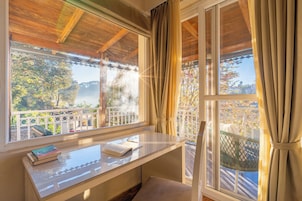 A room with a wooden ceiling, a window with curtains, a balcony with a railing, and a table with books and a notebook.