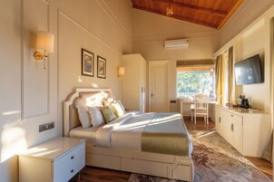 Un dormitorio con cama, mesitas de noche, escritorio, silla, televisor y ventana con vistas a árboles.