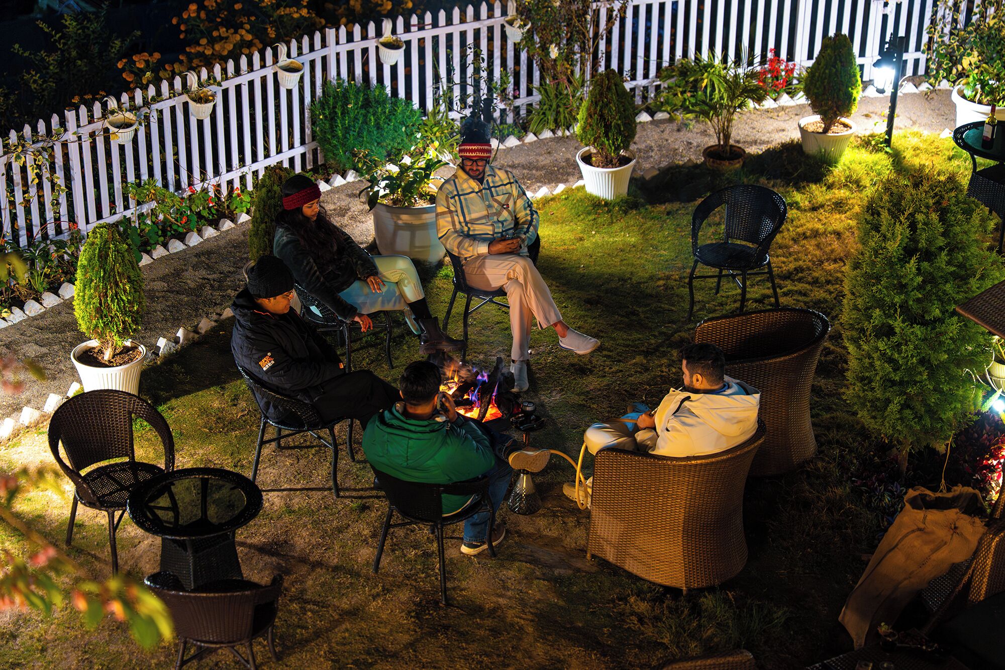An outdoor seating area with wicker chairs and tables, surrounded by potted plants and a white picket fence.