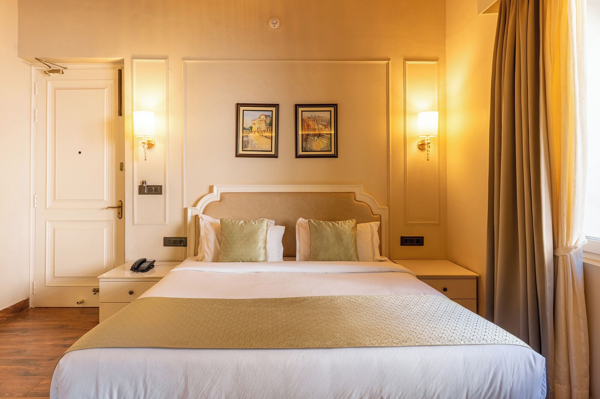 Habitación de hotel con una cama grande, dos mesitas de noche, una mesita auxiliar, una puerta y dos lámparas fijadas a la pared.