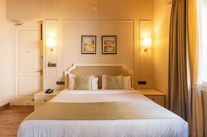 Habitación de hotel con una cama grande, dos mesitas de noche, una mesita auxiliar, una puerta y dos lámparas fijadas a la pared.