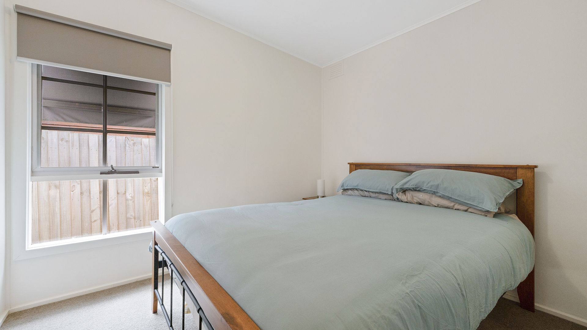 A bedroom with a single bed, a window with blinds, and a view of a wooden fence.
