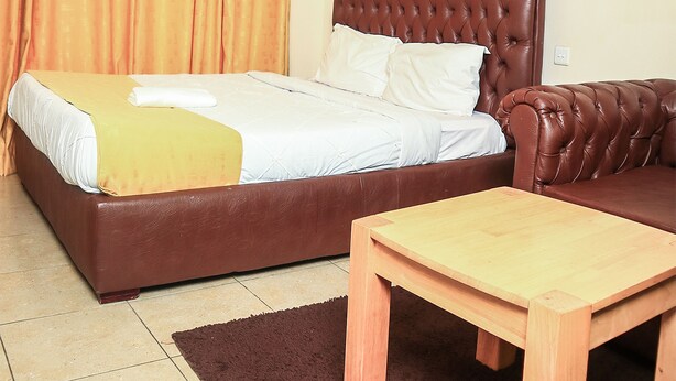 A bedroom with a large bed, a wooden nightstand, a brown leather sofa, and a ceiling light fixture. - West Suites Apartments (Nairobi)