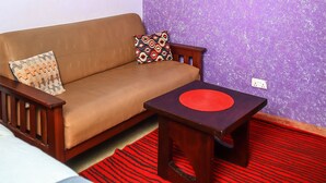 A room with a brown sofa, a small wooden table with a red circular object, a purple textured wall, and a ceiling light fixture. - West Suites Apartments (Nairobi)