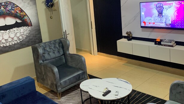 A modern living room with a grey sofa, a blue sofa, a white coffee table, and a TV on a black wall. - Magic City Apartment (Kasoa)