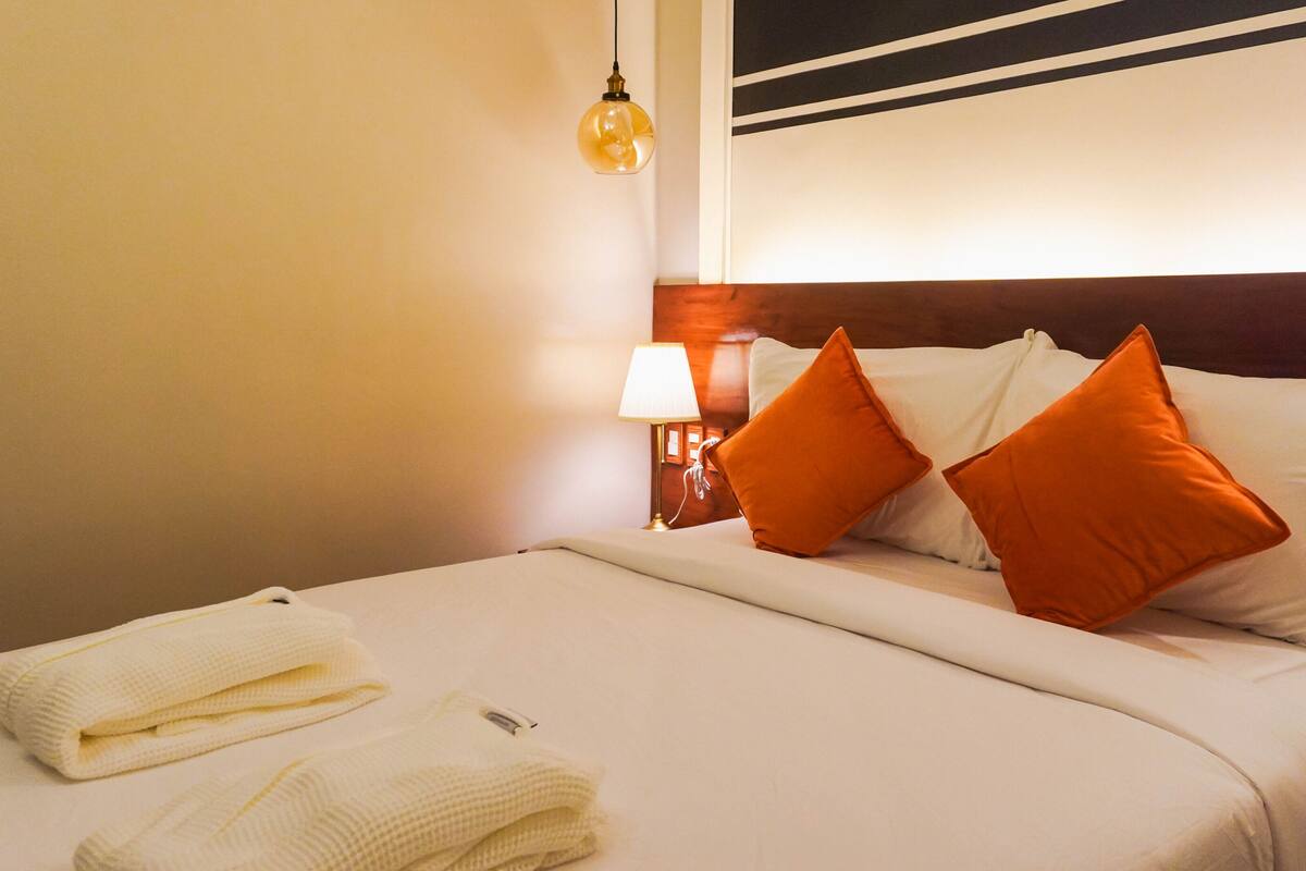 Een net opgemaakt bed met witte lakens en twee oranje kussens, een bedlamp en een muur met een zwart-wit streep patroon.