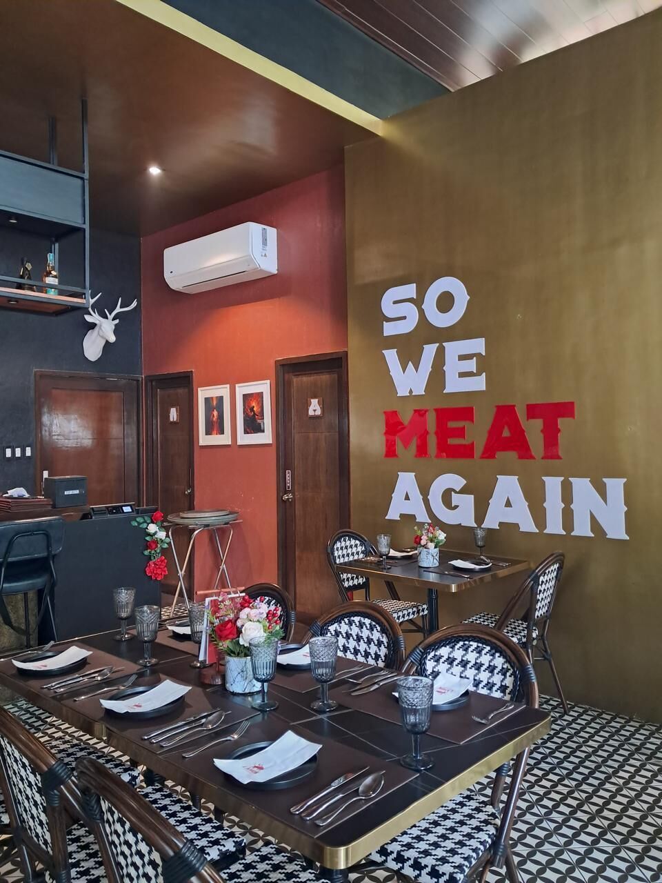 Interior de restaurante con una mesa para cuatro, una pared con el letrero 'SO WE MEAT AGAIN' y una cabeza de venado montada.