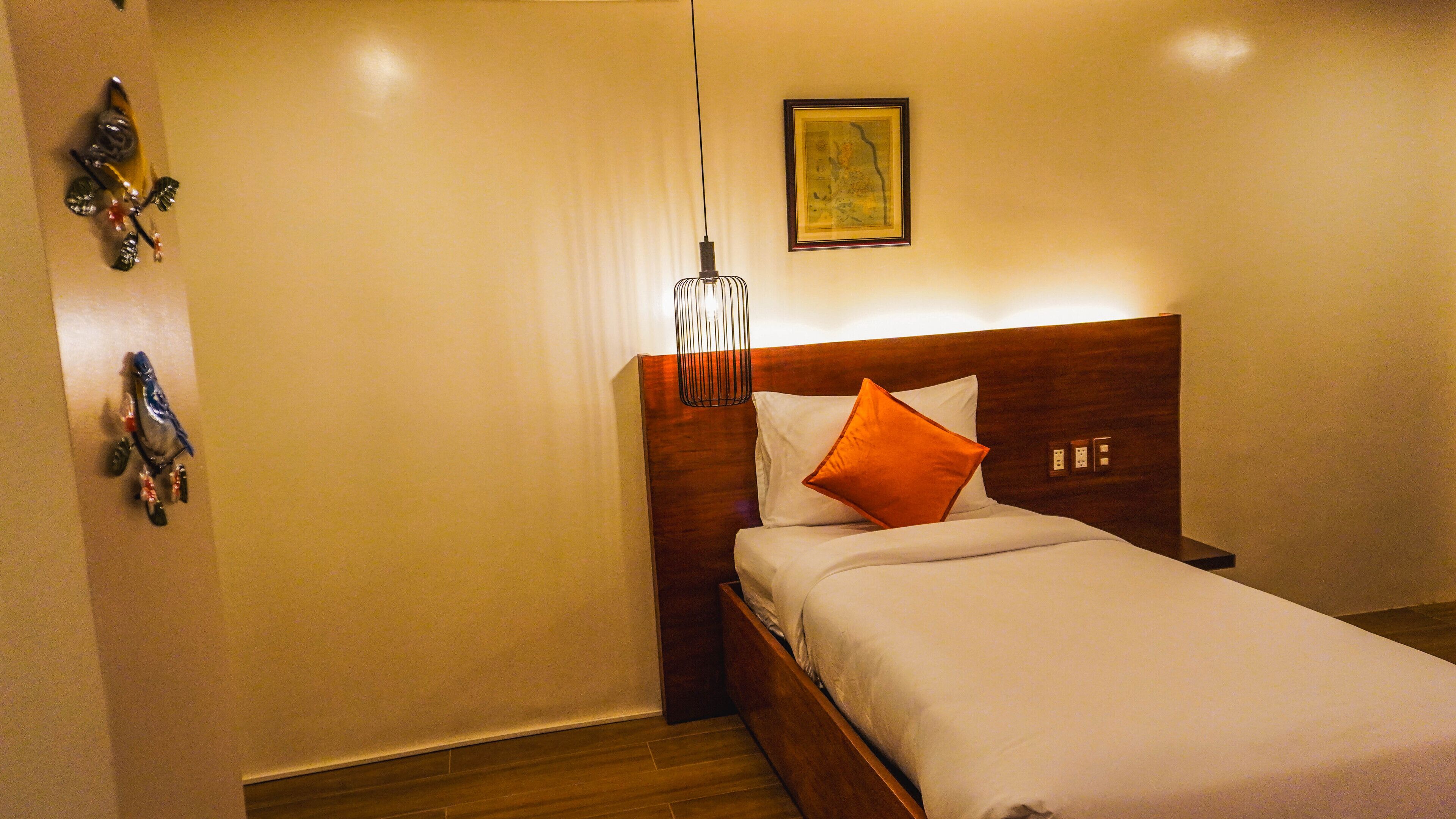Una cama bien tendida con ropa blanca y un almohadón naranja, cabecera de madera, lámpara colgante y un cuadro enmarcado en la pared.