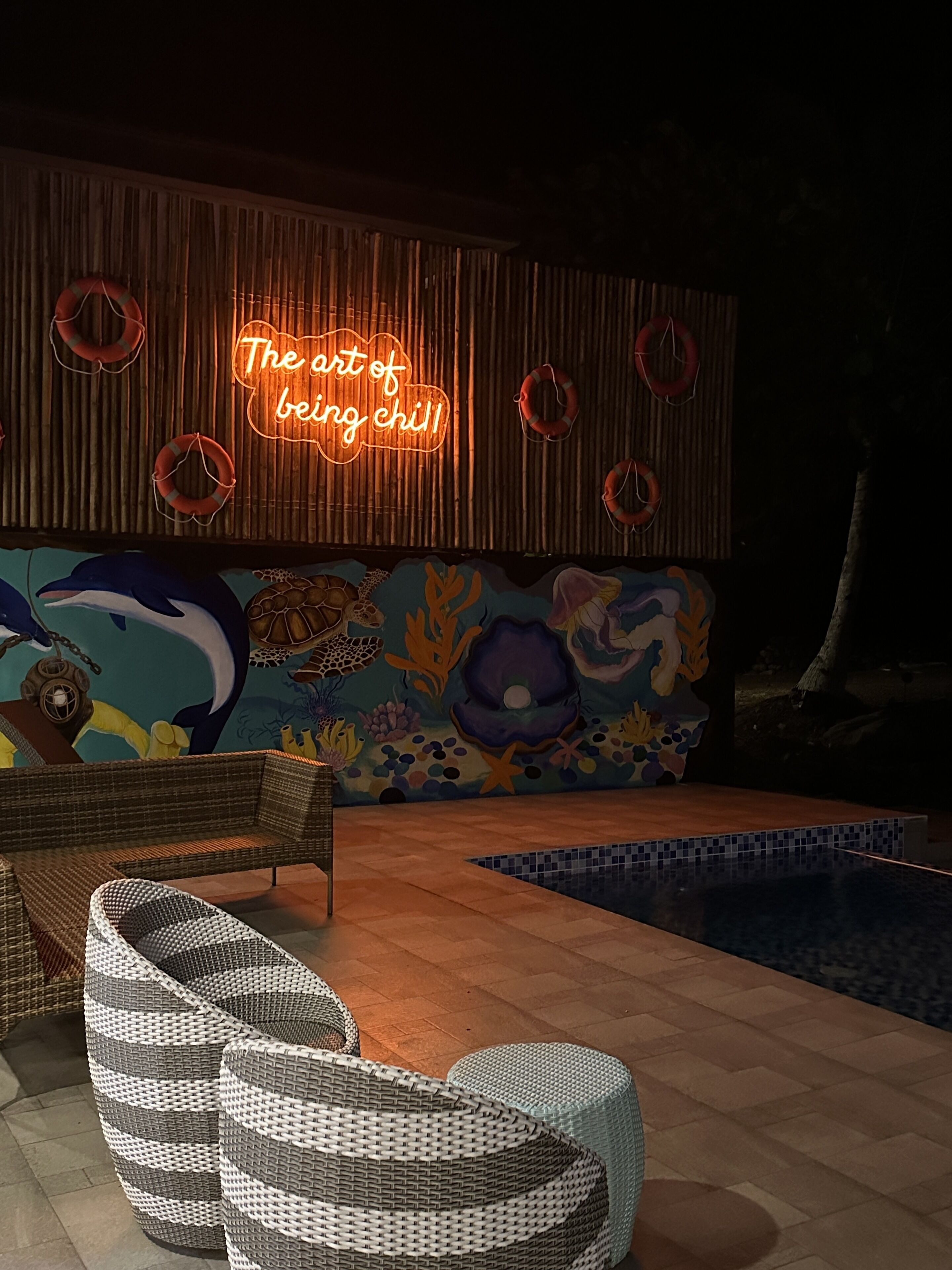 Zona de piscina con mobiliario de mimbre, un letrero de neón y un mural.