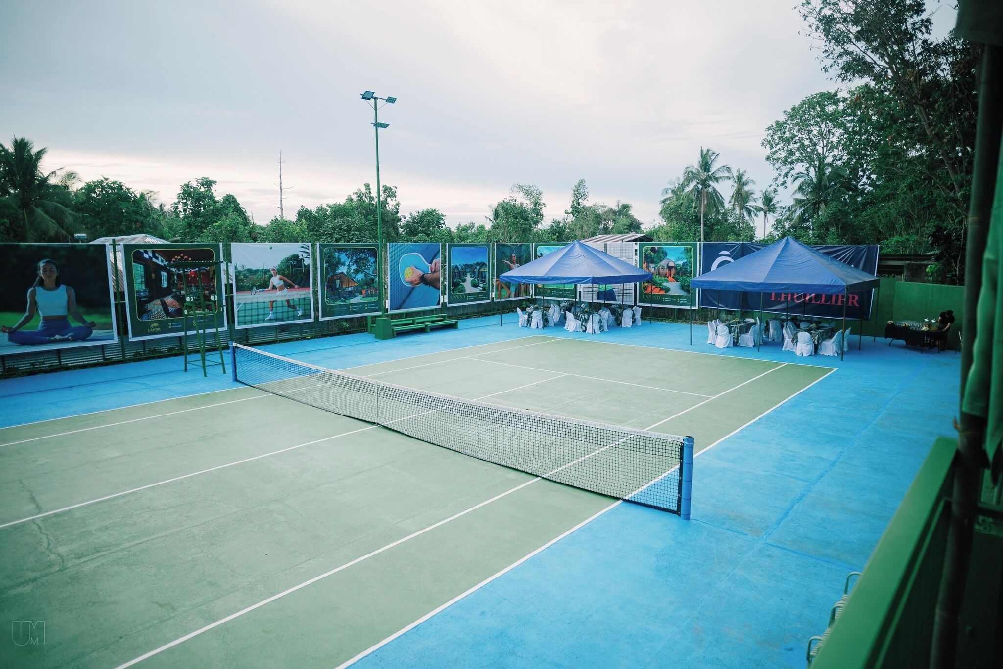 Cancha de tenis amb una xarxa, envoltada per una tanca, i una tenda amb el lema 'LIGUILIER'.